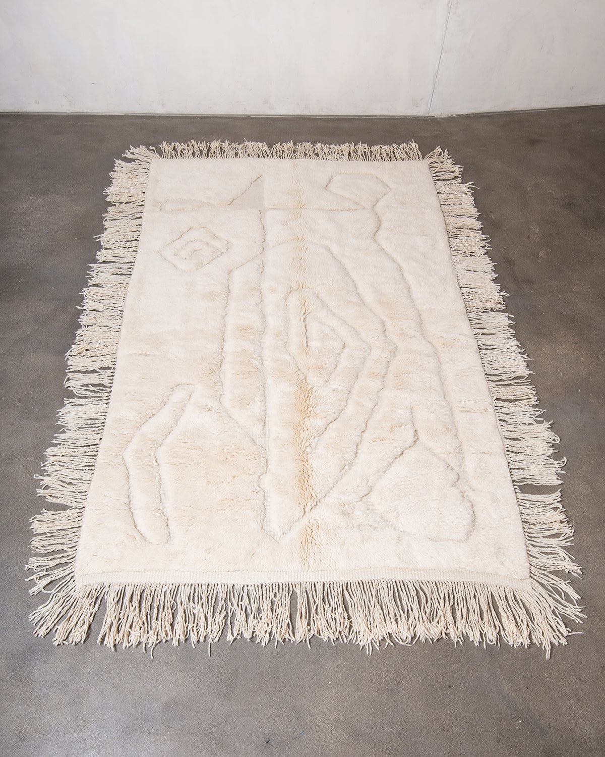 Berberlin x Malaika Raiss Beni M'rirt rug made of velvety cream sheep's wool with abstract woven design and natural wool tassels on all four sides, laying on concrete studio floor.